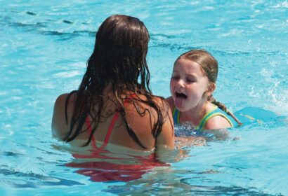 Two people are in a swimming pool, facing each other. One is a child sticking out their tongue, and the other is an adult with wet hair wearing a red swimsuit.