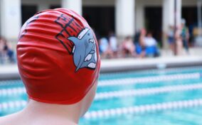 A swimmer wearing a red swim cap with a shark design stands poolside, observing the swimming pool.