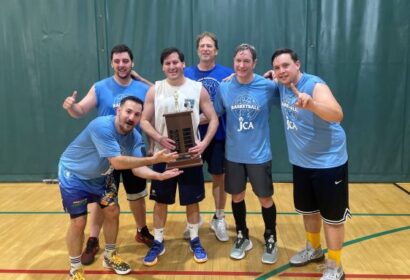 A group of six men in sportswear poses indoors on a basketball court, with one holding a trophy. They appear to be celebrating a victory.