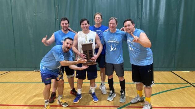 A group of six men in sportswear poses indoors on a basketball court, with one holding a trophy. They appear to be celebrating a victory.