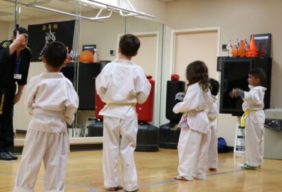 Children in white martial arts uniforms stand in formation, facing an instructor in black uniform in a dojo with mirrors on the wall.