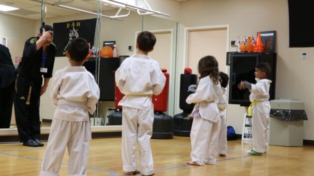 Children in white martial arts uniforms stand in formation, facing an instructor in black uniform in a dojo with mirrors on the wall.