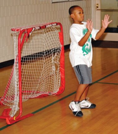 A child stands in front of a small indoor soccer goal with hands raised, wearing a white shirt, gray shorts, and sneakers on a gym floor.