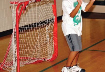 A child stands in front of a small indoor soccer goal with hands raised, wearing a white shirt, gray shorts, and sneakers on a gym floor.