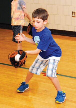 A young boy wearing a blue shirt and plaid shorts practices catching a baseball with a glove indoors.