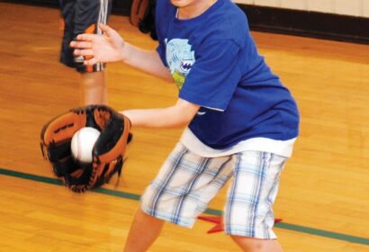 A young boy wearing a blue shirt and plaid shorts practices catching a baseball with a glove indoors.