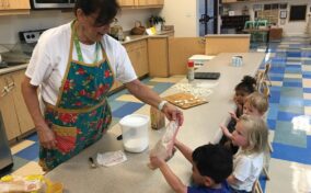 A woman in an apron helps a group of young children with a cooking activity in a classroom, passing a bag of ingredients to a child sitting at the table. Kitchen and classroom supplies are visible.