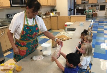 A woman in an apron helps a group of young children with a cooking activity in a classroom, passing a bag of ingredients to a child sitting at the table. Kitchen and classroom supplies are visible.