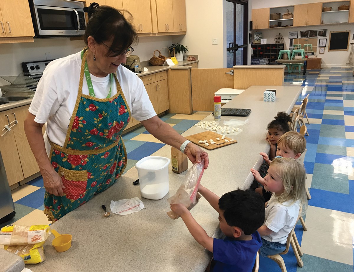 A woman in an apron helps a group of young children with a cooking activity in a classroom, passing a bag of ingredients to a child sitting at the table. Kitchen and classroom supplies are visible.