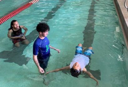 A swim instructor in a blue shirt is with a child who is floating on their back in a pool. Another person is observing nearby.