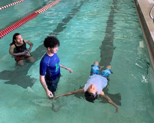 A swim instructor in a blue shirt is with a child who is floating on their back in a pool. Another person is observing nearby.