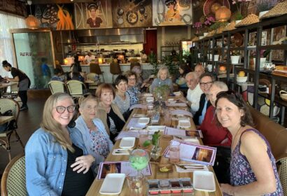 A group of eleven adults are seated around a long table in a restaurant, smiling at the camera. Menus and drinks are on the table, and the background displays the kitchen and restaurant decor.