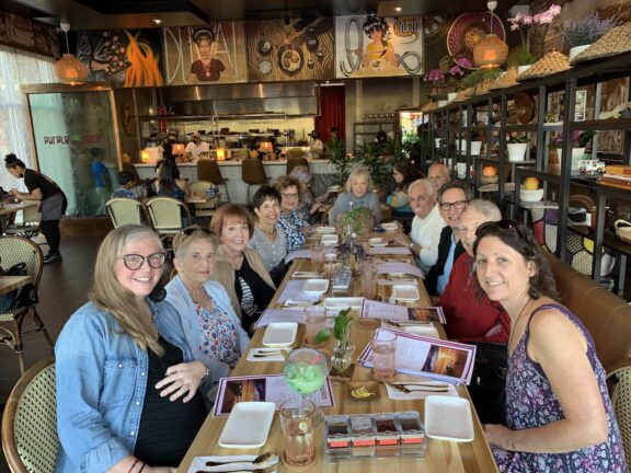 A group of eleven adults are seated around a long table in a restaurant, smiling at the camera. Menus and drinks are on the table, and the background displays the kitchen and restaurant decor.