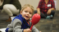 A young child wearing a blue and gray jacket smiles while holding a red maraca in a classroom. An adult and another child are blurred in the background.