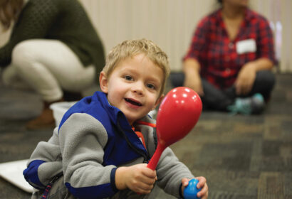 A young child wearing a blue and gray jacket smiles while holding a red maraca in a classroom. An adult and another child are blurred in the background.