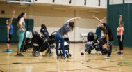 A group of adults, mostly women, stretches in a gymnasium while standing next to strollers with babies.