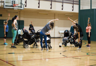 A group of adults, mostly women, stretches in a gymnasium while standing next to strollers with babies.