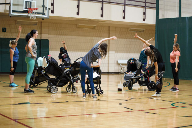 A group of adults, mostly women, stretches in a gymnasium while standing next to strollers with babies.