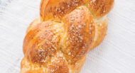 A loaf of braided bread with a golden crust on a white woven cloth, placed on a blue wooden surface.