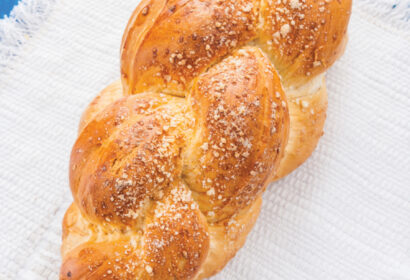 A loaf of braided bread with a golden crust on a white woven cloth, placed on a blue wooden surface.