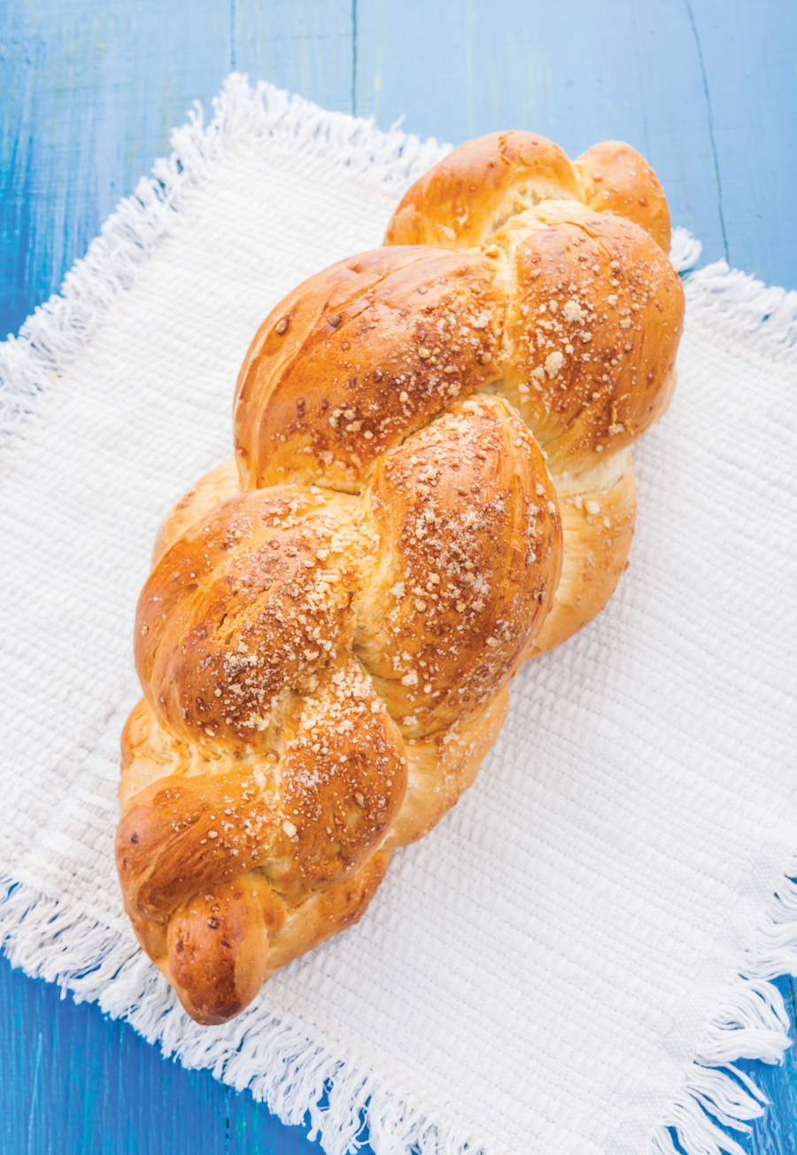 A loaf of braided bread with a golden crust on a white woven cloth, placed on a blue wooden surface.