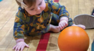 A baby in a patterned outfit is crawling on a gym floor, reaching for an orange ball in front of them.