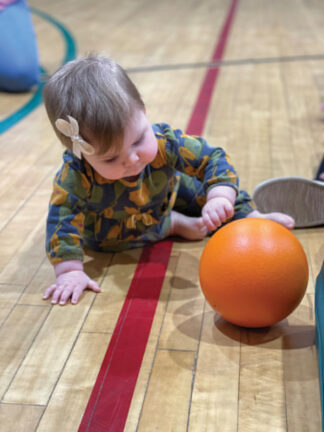 A baby in a patterned outfit is crawling on a gym floor, reaching for an orange ball in front of them.