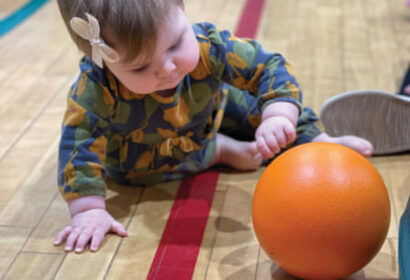 A baby in a patterned outfit is crawling on a gym floor, reaching for an orange ball in front of them.