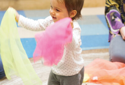 A toddler in a white shirt and striped pants holds colorful scarves while smiling in a room with shelves and mats.