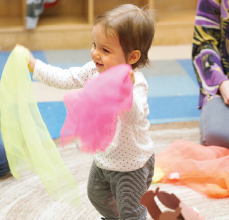 A toddler in a white shirt and striped pants holds colorful scarves while smiling in a room with shelves and mats.