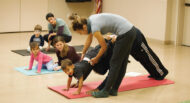 Instructor assisting a child in a yoga pose on a mat while other children and adults sit or perform yoga in the background in a classroom setting.