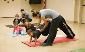 Instructor assisting a child in a yoga pose on a mat while other children and adults sit or perform yoga in the background in a classroom setting.