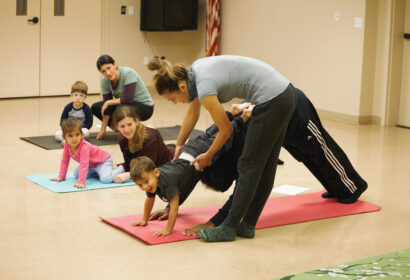 Instructor assisting a child in a yoga pose on a mat while other children and adults sit or perform yoga in the background in a classroom setting.