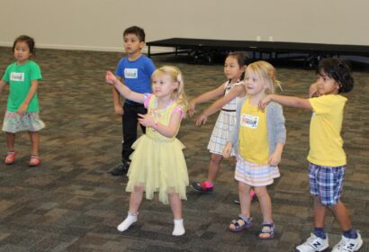 Six children stand in a room, some pointing or gesturing, in a playful or interactive activity. The floor is carpeted, and a riser is in the background.