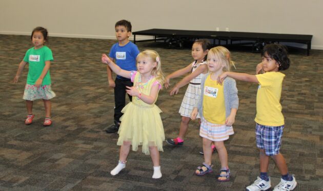 Six children stand in a room, some pointing or gesturing, in a playful or interactive activity. The floor is carpeted, and a riser is in the background.