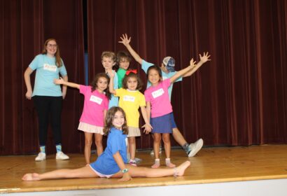 A group of children and one adult stand on a stage, with some children making energetic poses and one child performing the splits in the front. They are wearing colorful shirts and name tags.