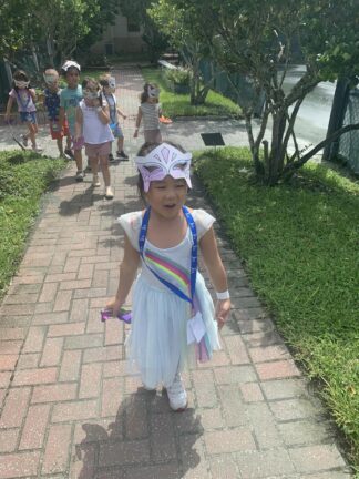 A young child wearing a butterfly mask and rainbow dress walks on a brick path followed by a group of children outdoors.