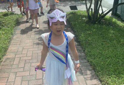 A young child wearing a butterfly mask and rainbow dress walks on a brick path followed by a group of children outdoors.