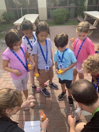 Several children standing on a patio, all wearing blue lanyards, closely observe as two adults demonstrate a small science experiment on the ground.