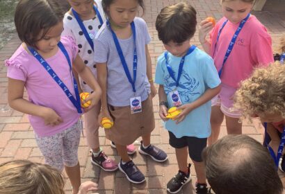 Several children standing on a patio, all wearing blue lanyards, closely observe as two adults demonstrate a small science experiment on the ground.