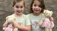 Two young girls stand side by side, each holding a teddy bear dressed in a pink ballet outfit. Both girls are smiling and wearing matching light-colored shirts with a similar teddy bear print.