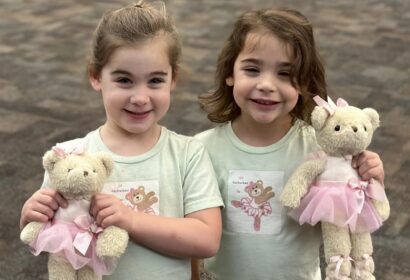 Two young girls stand side by side, each holding a teddy bear dressed in a pink ballet outfit. Both girls are smiling and wearing matching light-colored shirts with a similar teddy bear print.