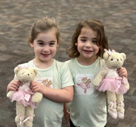 Two young girls stand side by side, each holding a teddy bear dressed in a pink ballet outfit. Both girls are smiling and wearing matching light-colored shirts with a similar teddy bear print.