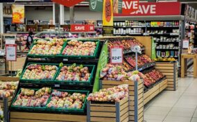 A supermarket produce section displaying various fresh fruits, including apples and pears, arranged in wooden crates. Sale signs and price tags are visible above the produce. Shelves with other products are in the background.