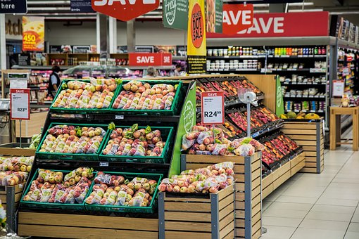 A supermarket produce section displaying various fresh fruits, including apples and pears, arranged in wooden crates. Sale signs and price tags are visible above the produce. Shelves with other products are in the background.