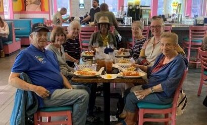 A group of seven older adults sitting at a restaurant table, sharing a meal with plates of food in front of them, inside a brightly decorated eatery with pink chairs and teal booths.