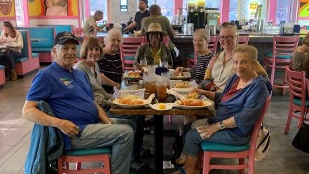 A group of seven older adults sitting at a restaurant table, sharing a meal with plates of food in front of them, inside a brightly decorated eatery with pink chairs and teal booths.