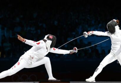 Two fencers in protective gear compete in a match, both brandishing their epees. One fencer lunges forward while the other parries, both connected by the scoring cord in an arena setting.