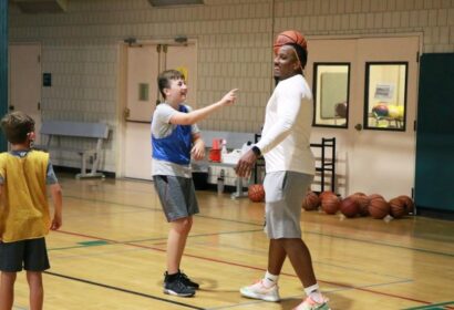 A man with a basketball balanced on his head stands in a gym while smiling at a pointing child, with another child looking on.