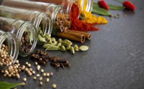 Various spices, including cinnamon sticks, cardamom pods, cloves, bay leaves, and chili peppers, spilling from glass jars onto a gray surface.